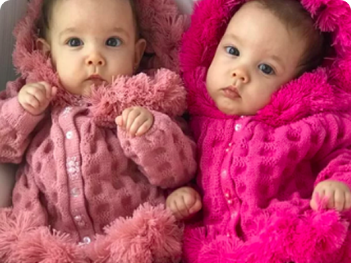 Duas bebês gêmeas vestindo macacões felpudos rosa e fúcsia, deitadas lado a lado.